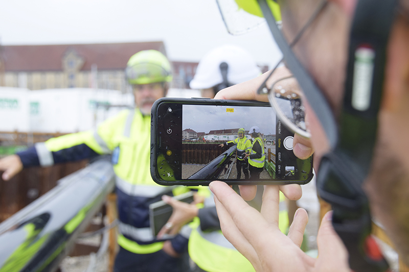 Papirøen - Christiansholm Ø. Mobil tager billede af 2 der snakker ved kanten af byggegruben.